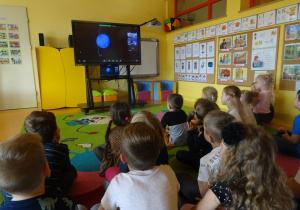 Grupa dzieci siedzi przed ekranem i z zaciekawieniem ogląda poszczególne planety.
